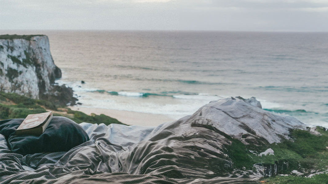 Das Bild zeigt kuschelige Bettwäsche an einem Strand mit Felsen. Es liegt ein Buch auf der Bettwaesche und in der Ferne sieht man das Meer.