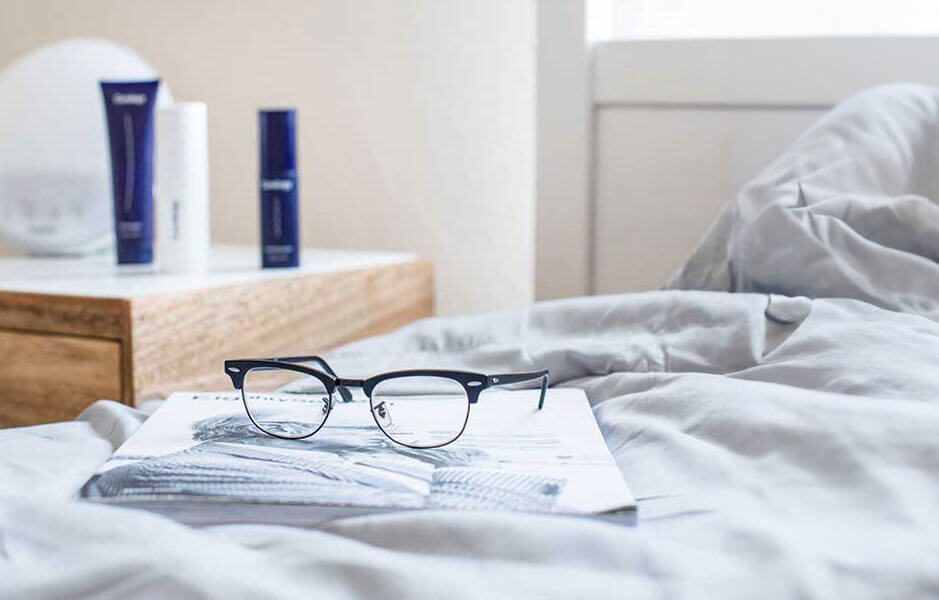 Zu sehen ist ein Bett mit weisser Bettwaesche, darauf liegend ein Buch und eine aufgeklappte Brille. Im Anschnitt ist eine Kommode zu sehen, auf der so etwas wie Pflegeprodukte stehen.